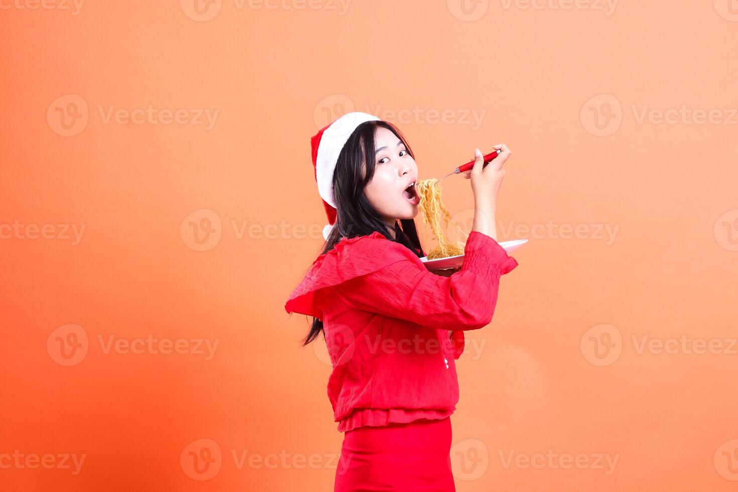expression of ladies wearing christmas dress, Santa hat, left slant focus eating noodles looking at camera, hand holding noodle fork and white plate filled with noodles, isolated on orange background photo