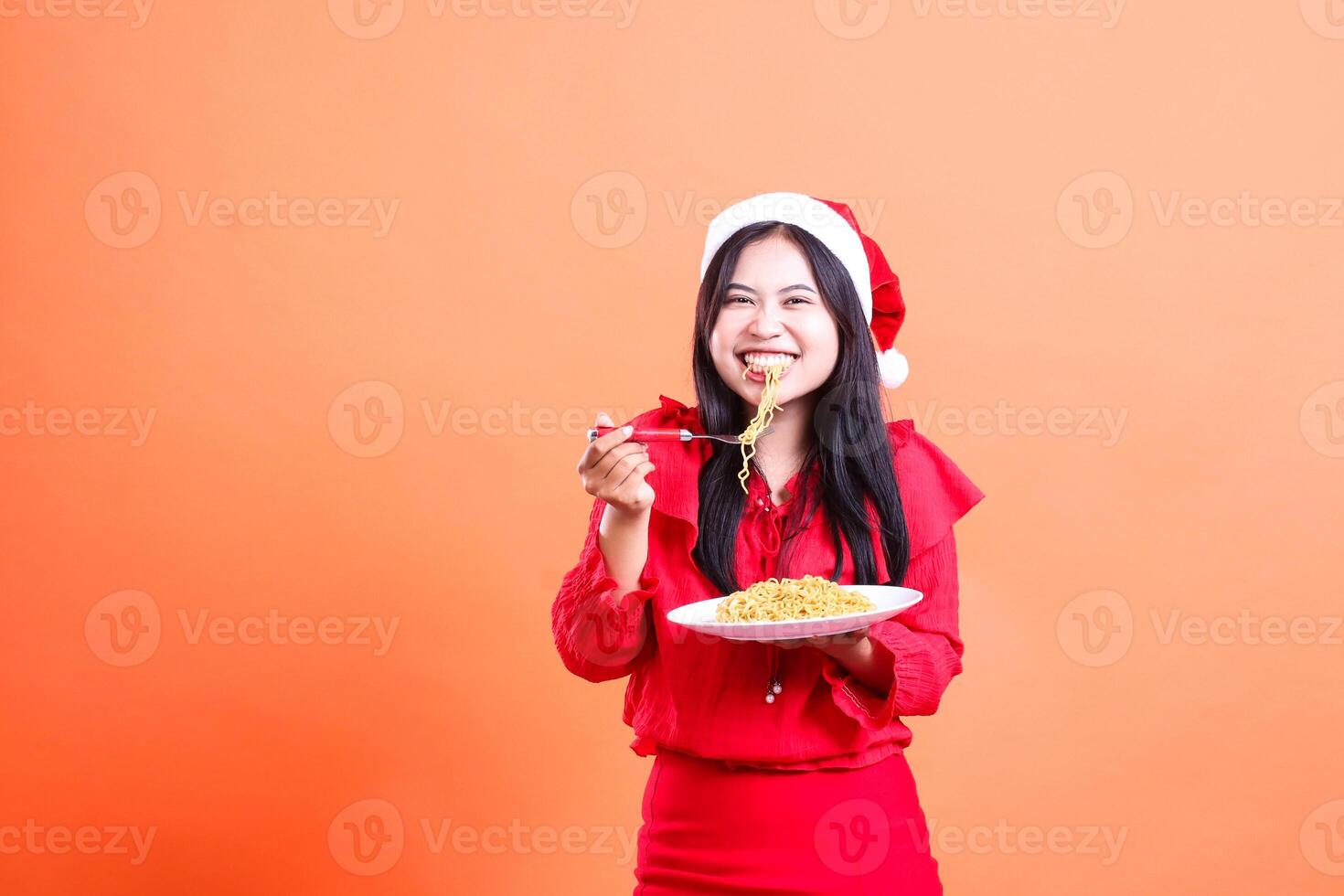 portrait of woman wearing christmas dress, santa hat, cheerful biting noodle, hand holding noodle fork on white plate celebrating christmas, isolated on orange background photo