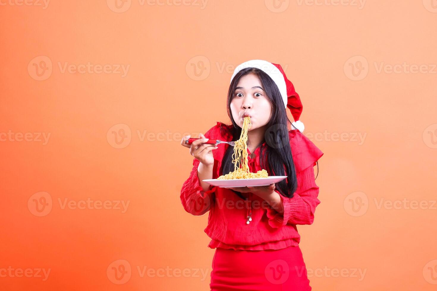 gesture of beautiful girl wearing red christmas blouse, Santa hat, surprised slurping noodles to camera, hand holding fork filled with noodles and white plate, isolated on orange background photo