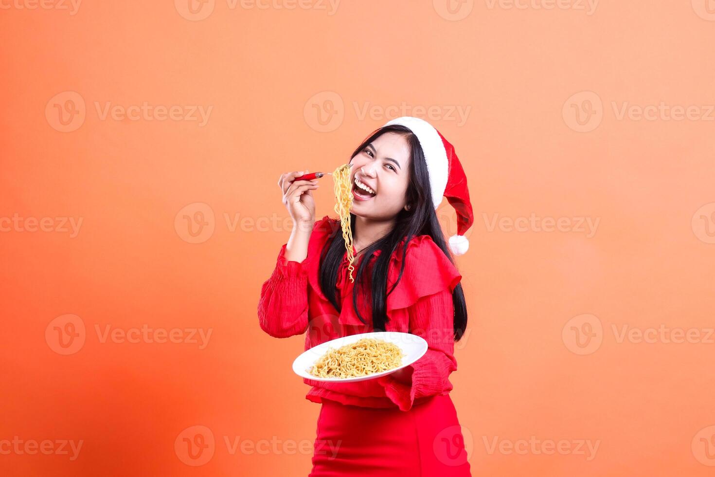 cheerful indonesia woman tilted front view, hand holding fork filled with noodles from white plate while showing off, wearing red christmas blouse, Santa hat, isolated on orange background photo