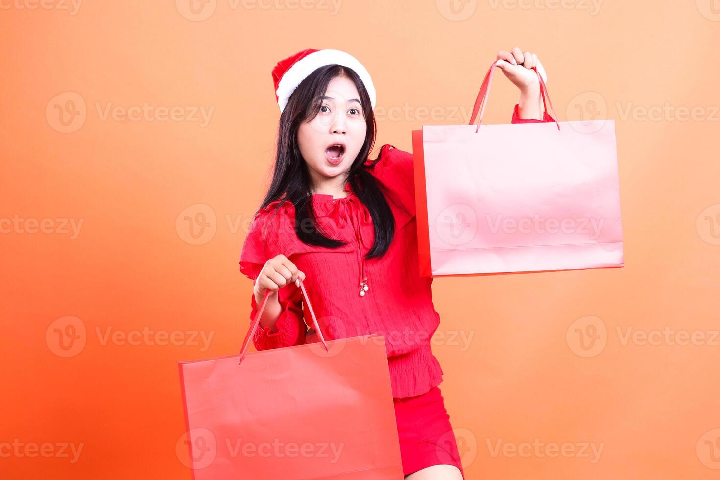 gesture of young girl standing wearing red blouse and santa hat, standing sideways left surprised to left both hands holding red bag of christmas gifts up and down, isolated on orange background photo