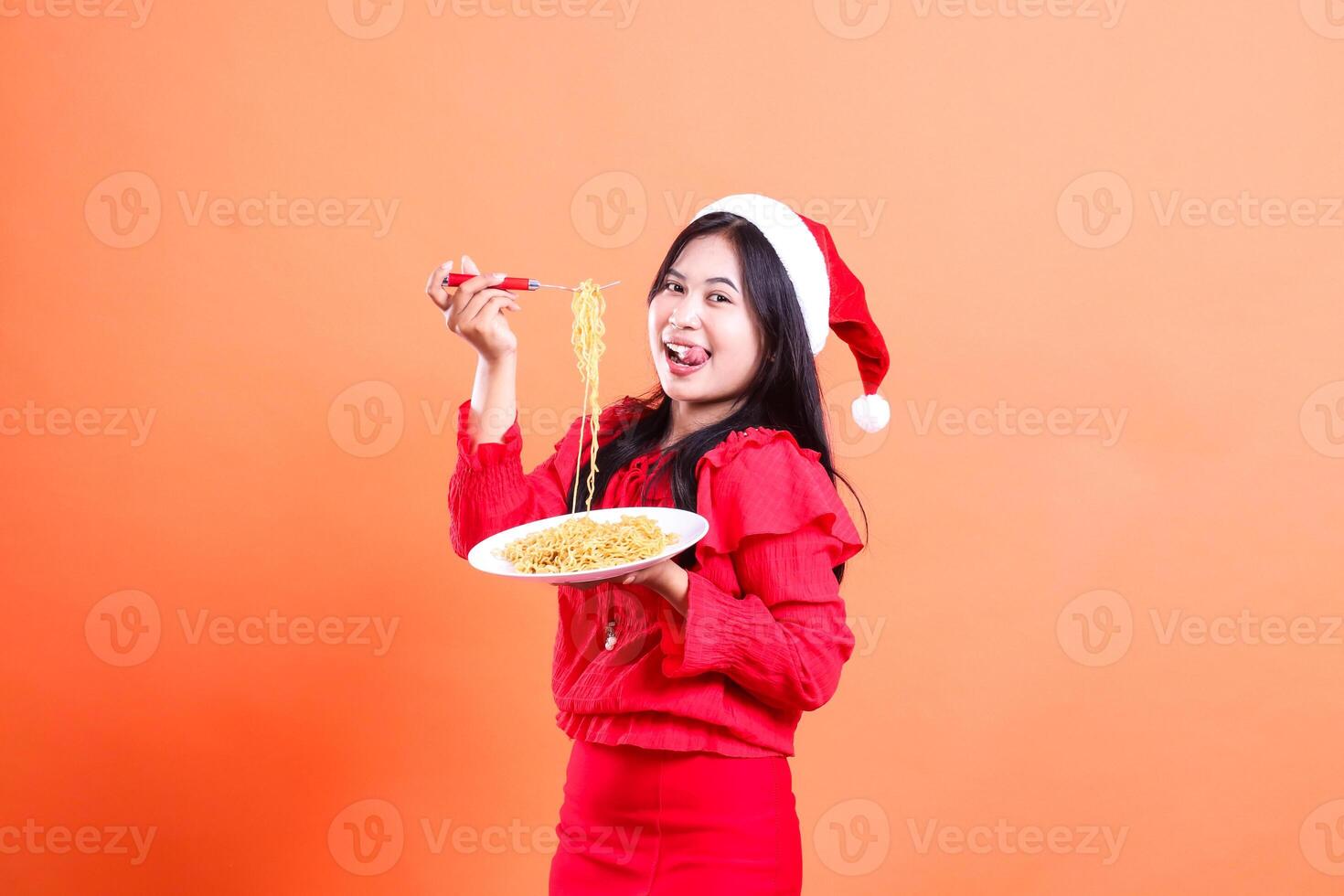 gesture of woman wearing red christmas blouse, Santa hat, happy hungry looking at camera, hand holding fork filled with noodles up and white plate filled with noodles, isolated on orange background photo