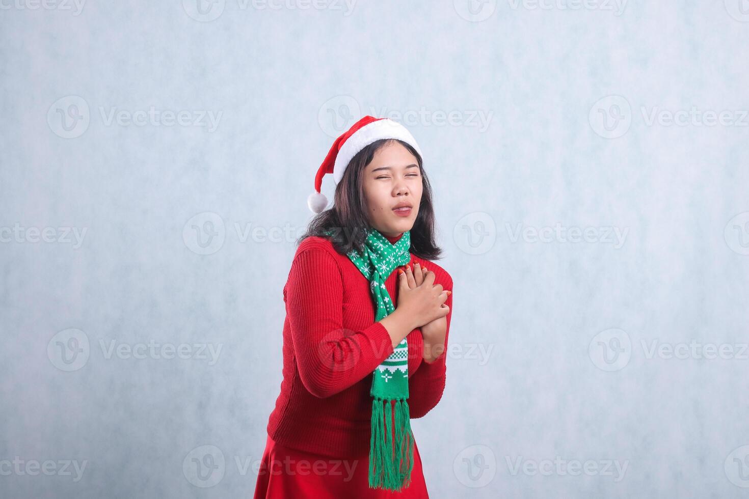 charm of young indonesia woman wearing red christmas sweater, santa hat and scarf, left side pain heart pain close eyes out tongue both hands hold heart, isolated on white background photo