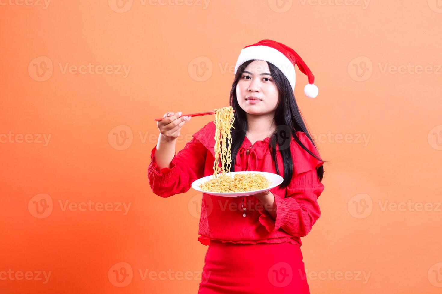 portrait of a women wearing Christmas clothes, Santa hat, elegantly looking at the camera, hands holding chopsticks and white plate filled with noodles ready to eat, isolated on orange background photo