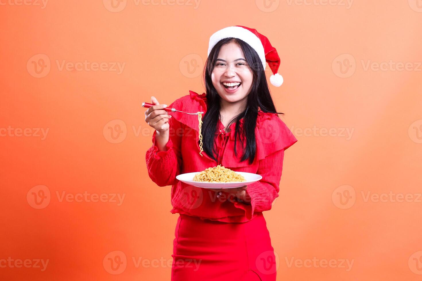portrait of woman wearing christmas blouse, Santa hat, cheerful to camera, hand holding fork filled with little noodles and noodles in white plate celebrating christmas, isolated on orange background photo