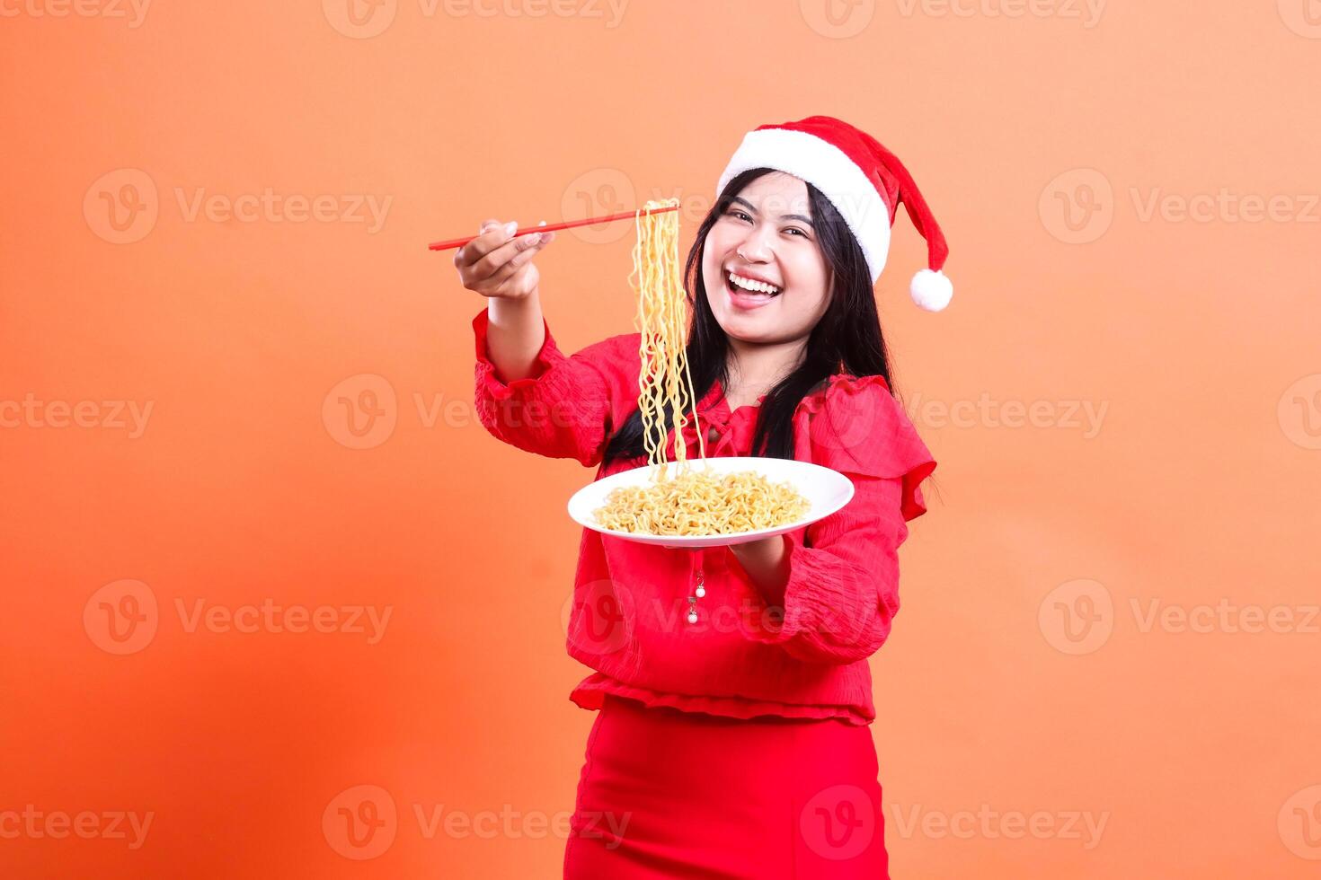 portrait of female wearing blouse, Santa hat, cheerful looking forward, hands holding chopsticks filled with noodles upwards and white plate filled with noodles looking forward, isolated orange photo