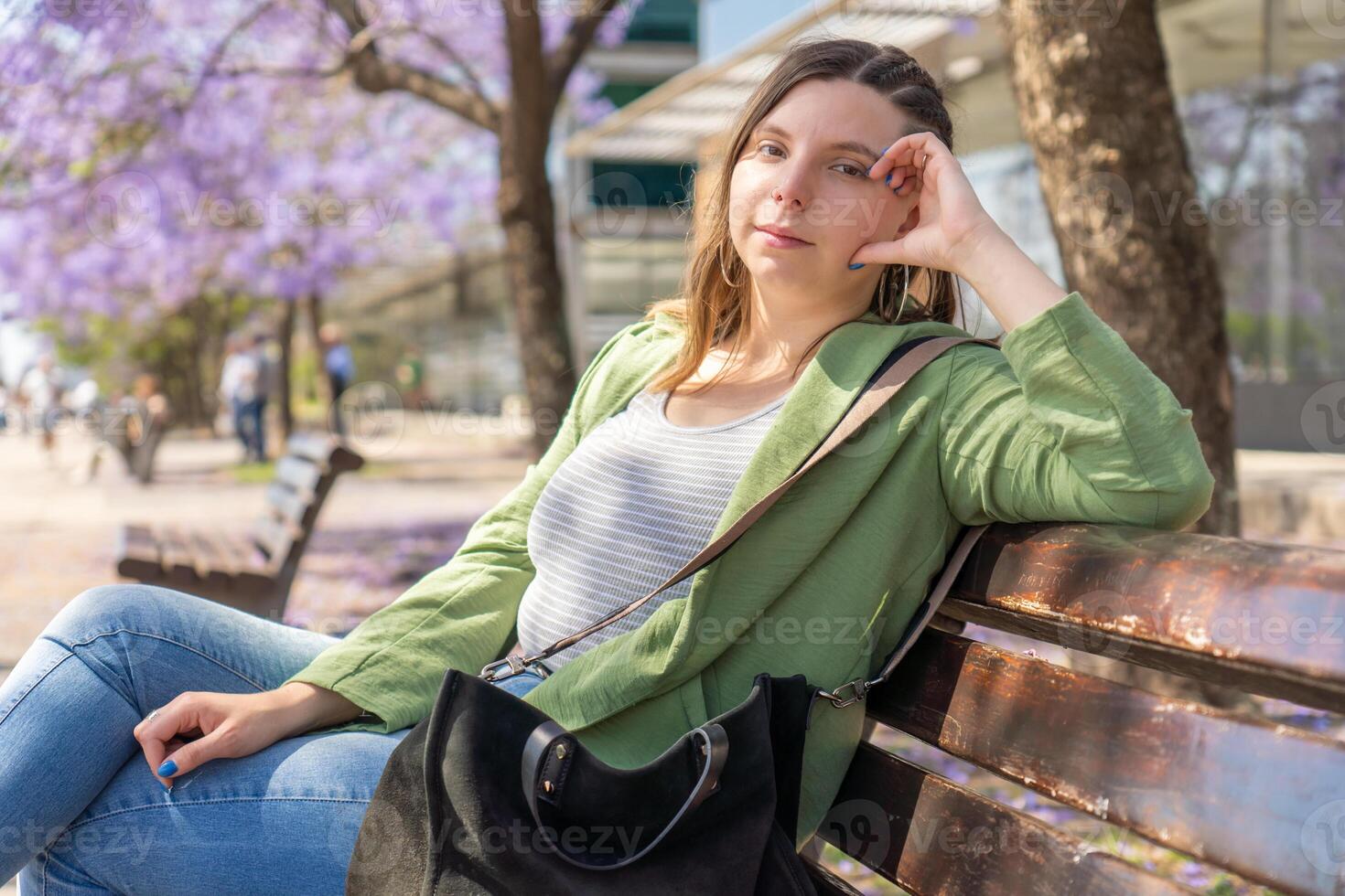 Woman looking at the camera while relaxing on a bench in a sunny park with blooming purple trees photo