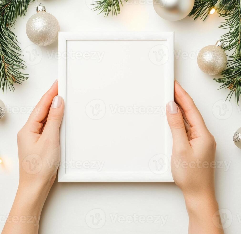 blank white frame mockup held by hands, surrounded by festive Christmas decorations like ornaments and pine branches, creating cheerful holiday atmosphere photo
