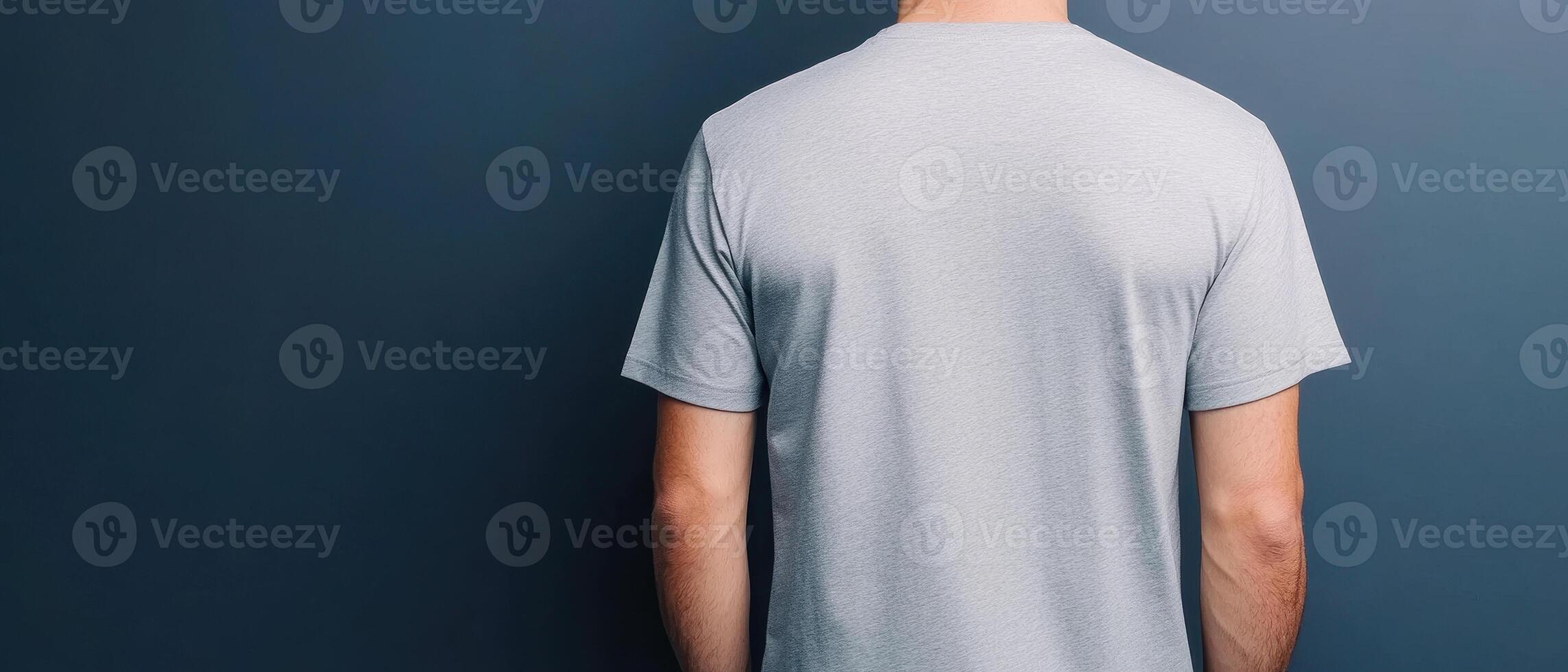 man wearing grey t shirt stands against dark blue wall, showcasing back view of shirt. simple design highlights fabric texture and fit, creating casual and modern look photo