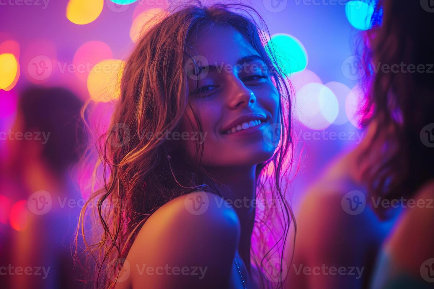 A young woman smiles joyfully at a lively gathering filled with colorful lights and energetic people around her, creating a festive atmosphere. photo