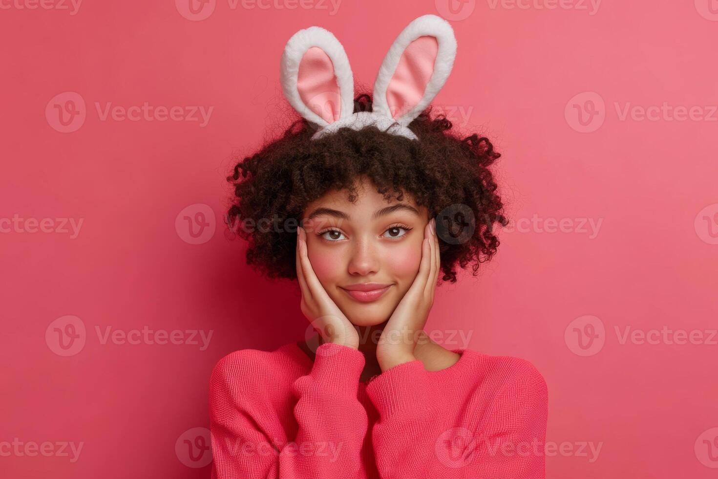 Young woman wearing bunny ears and a pink sweater poses playfully against a bright pink background, showcasing a fun expression of joy photo