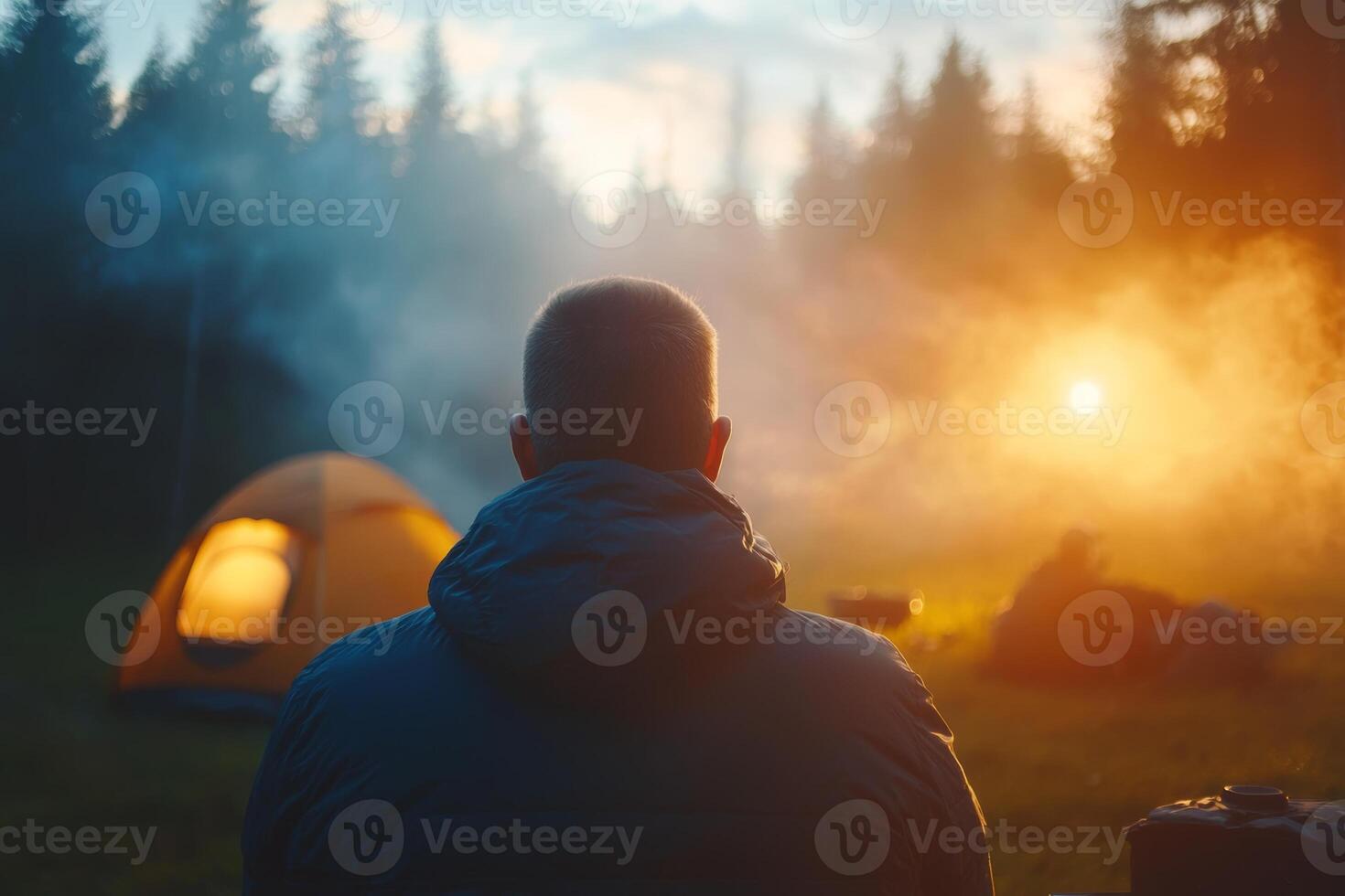 A camper gazes at the glowing campfire while enjoying a tranquil evening beside a tent in the forest during sunset photo