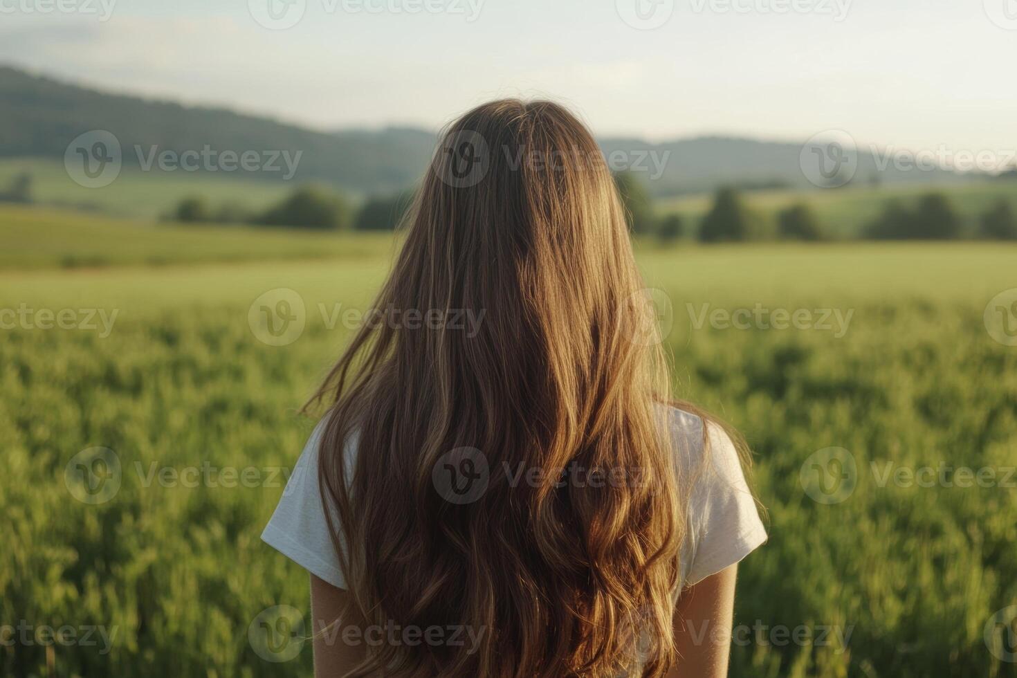 A young woman with long hair stands alone in a green field, gazing at the distant hills on a bright sunny afternoon photo