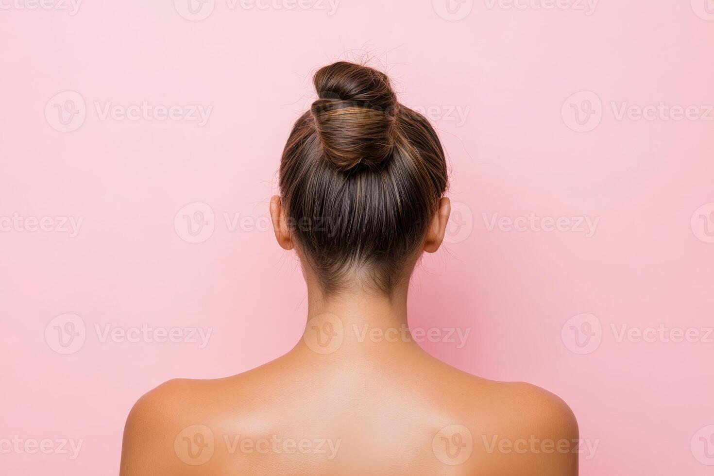 A woman with a sleek bun hairstyle displays her back against a soft pink background in a minimalist setting, highlighting hair styling elegance photo