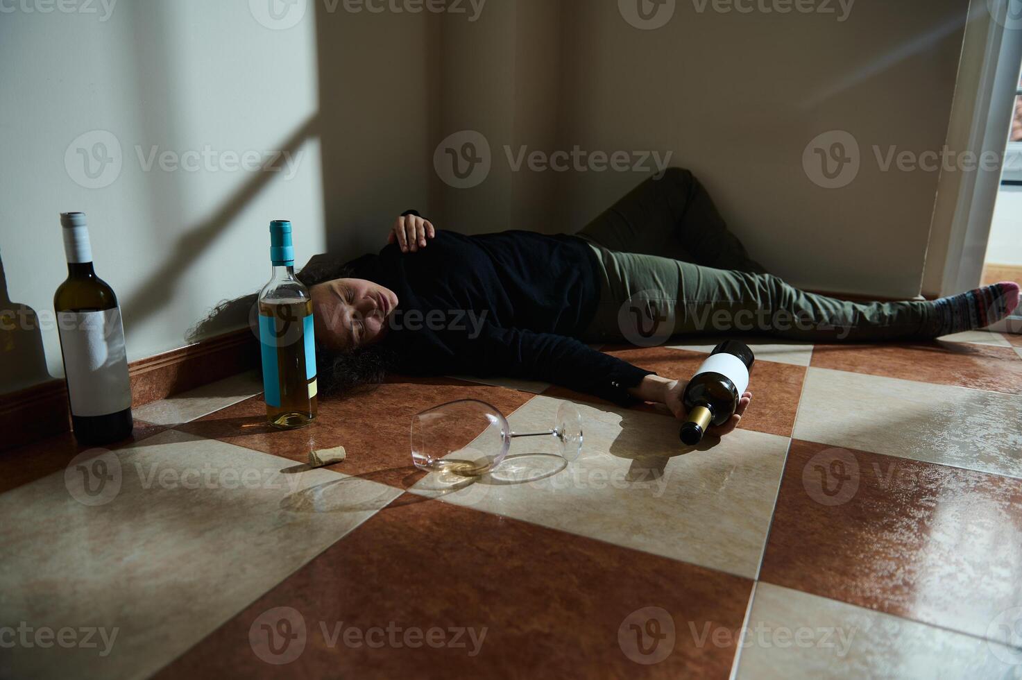 Person on tiled floor surrounded by empty alcohol bottles with glassware photo