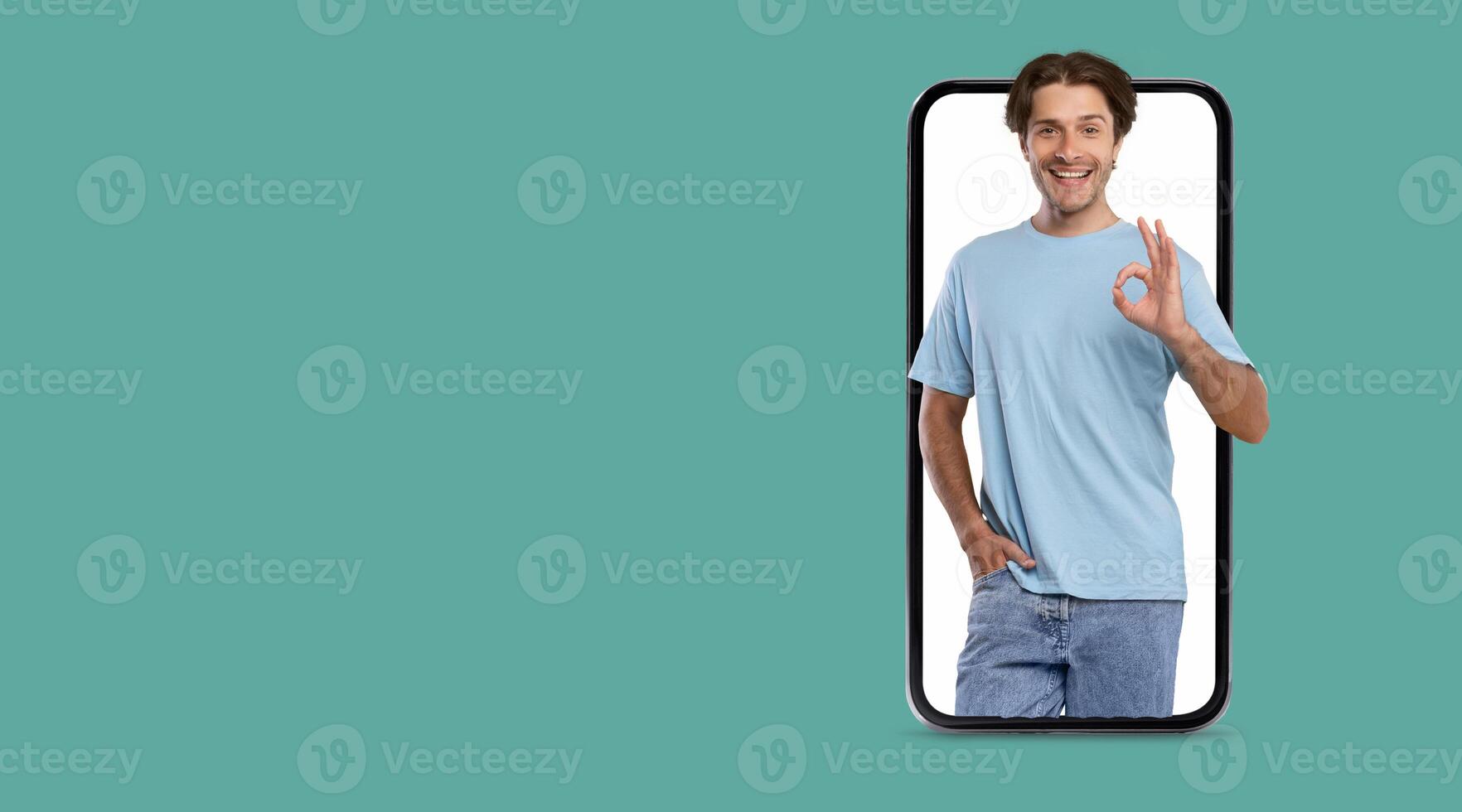 A young individual wearing a light blue shirt stands confidently while displaying an okay gesture. The large smartphone screen emphasizes their friendly demeanor against a clean background. photo