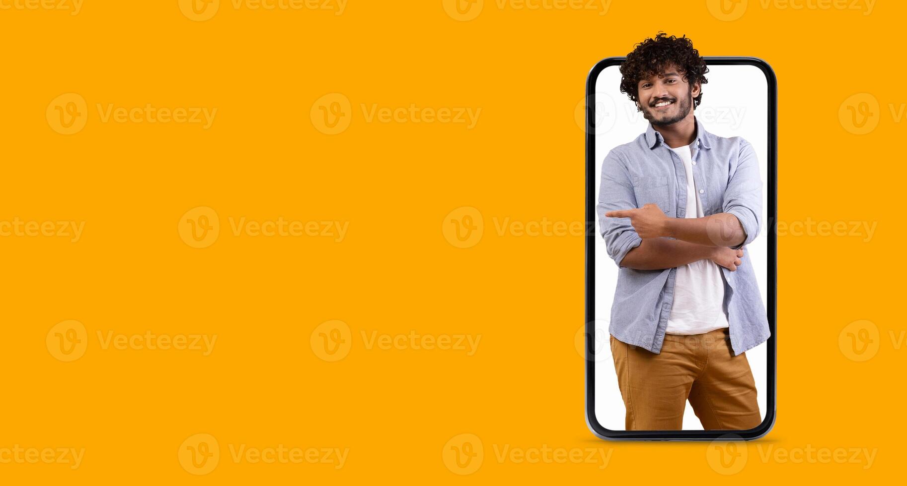 A cheerful young man smiles and poses with crossed arms while appearing to interact with a large smartphone screen in a vibrant yellow background. The scene focuses on modern communication. photo