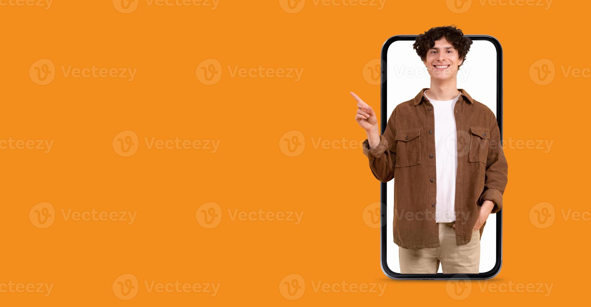 A young person stands in front of a large smartphone with a bright orange background. They are smiling and pointing, engaging in online communication or social media interaction. photo