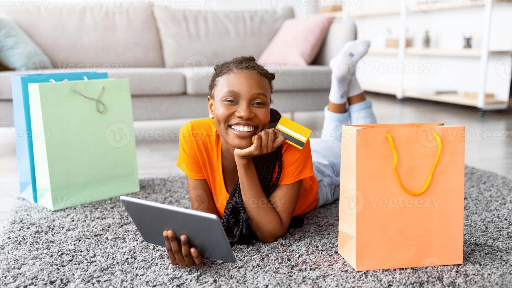 Online sales, discounts, e-commerce concept. Positive black woman lying on floor with tablet and credit card, shopping on internet from home. Cheery Afro lady with gift bags purchasing in web store photo