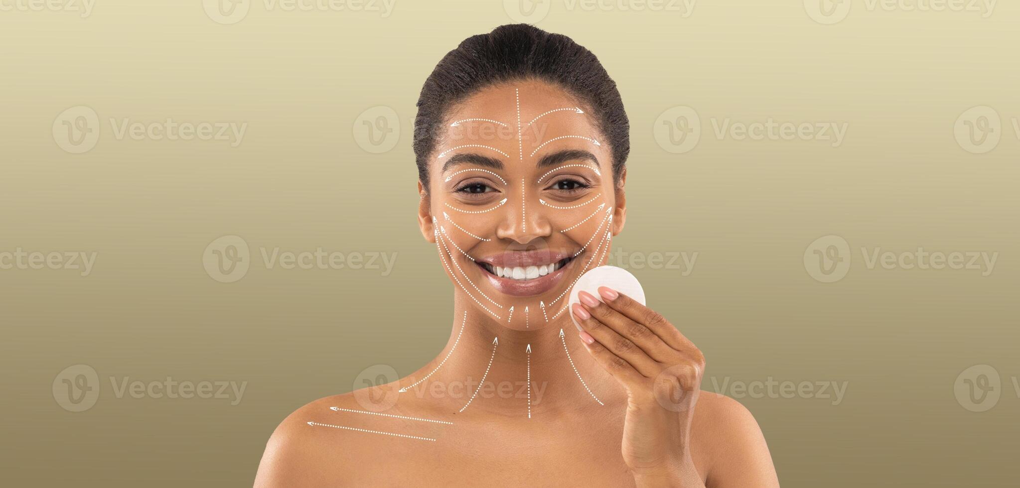 African American woman is engaged in her beauty routine, smiling while applying skin care products. She is in a calm, isolated setting with a neutral background, emphasizing self-care and pampering. photo