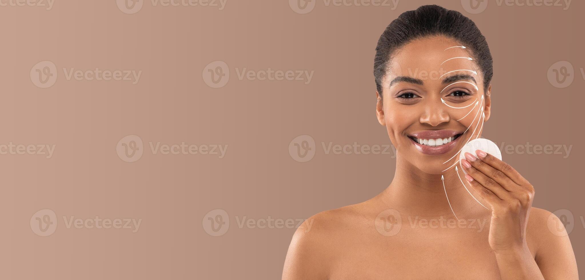 African American woman engages in her skincare routine, applying a product to her face while smiling. She is in a minimalist setting with soft lighting, emphasizing relaxation and self-care. photo