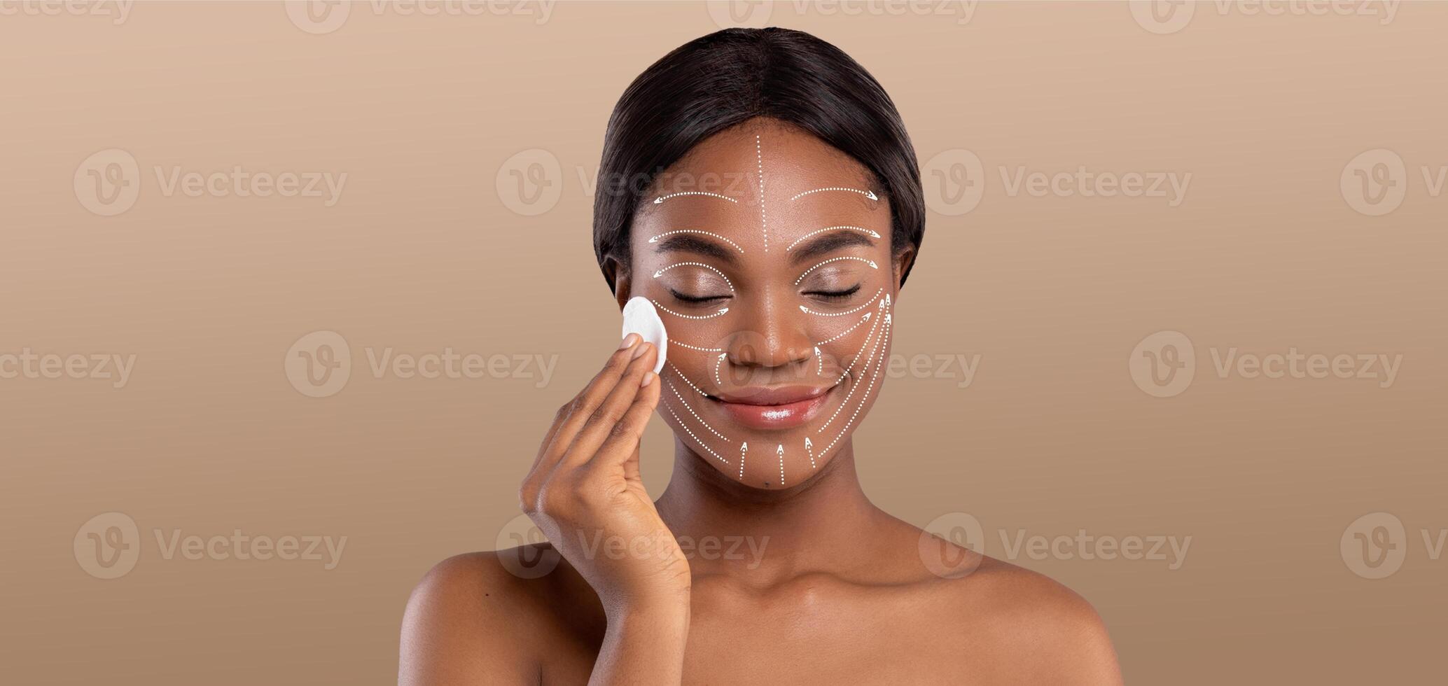 African American woman engages in a relaxing skin care routine, applying a product to her face with a gentle smile. She is isolated against a neutral background, highlighting her pampering moment. photo