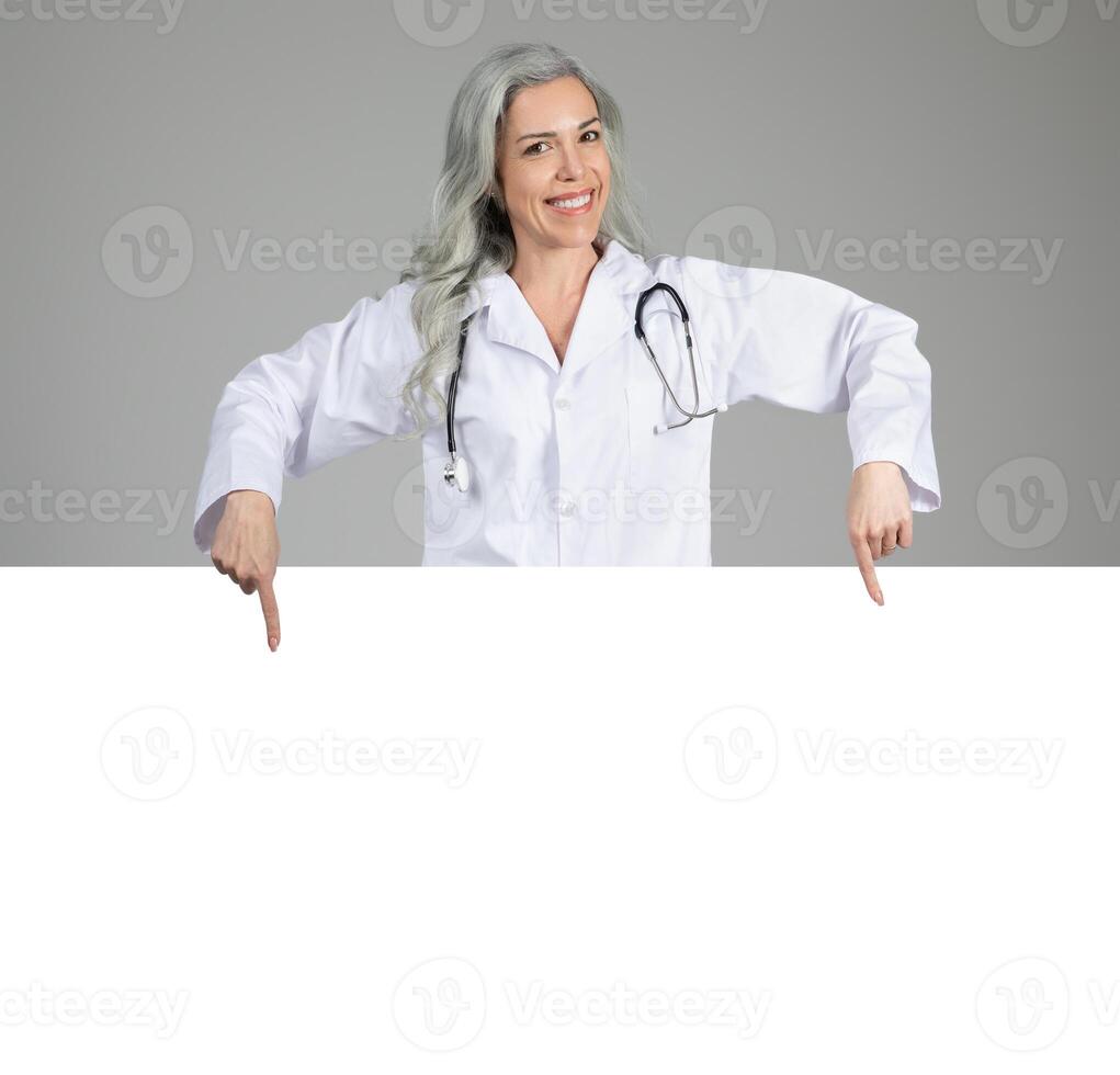 Middle aged woman in a doctor's coat smiles while pointing downwards, suggesting readiness to share important information about healthcare. She is isolated against a neutral background. photo