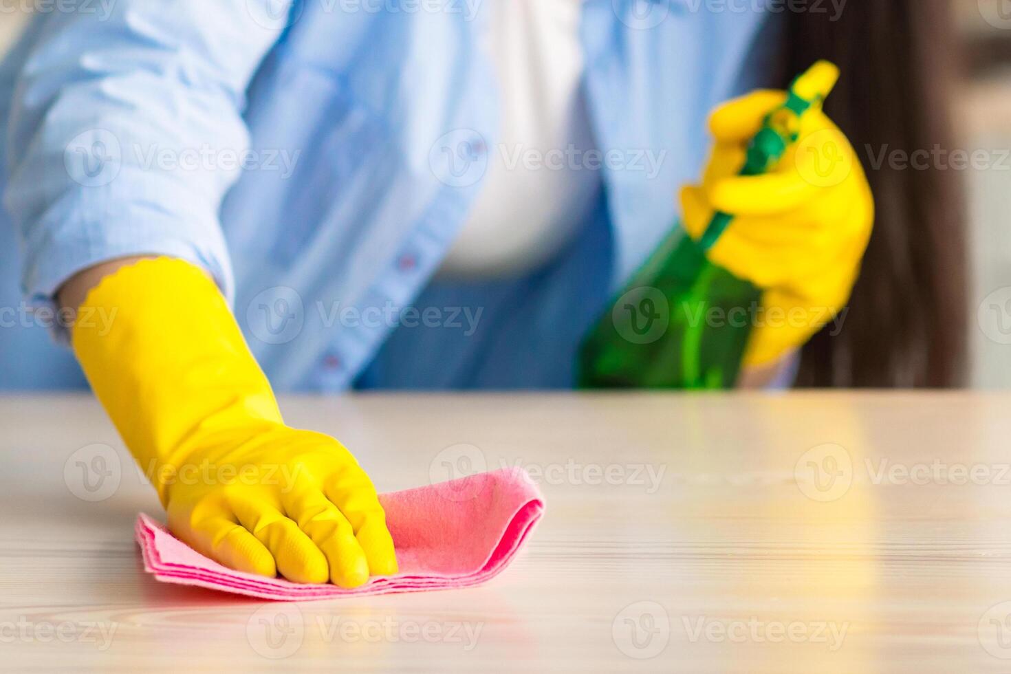 Spring Cleaning Concept. Closeup of unrecognizable young woman wearing protecitve yellow rubber gloves polishing table, holding spray bottle using microfiber rag, wiping dust, selective focus on hand photo