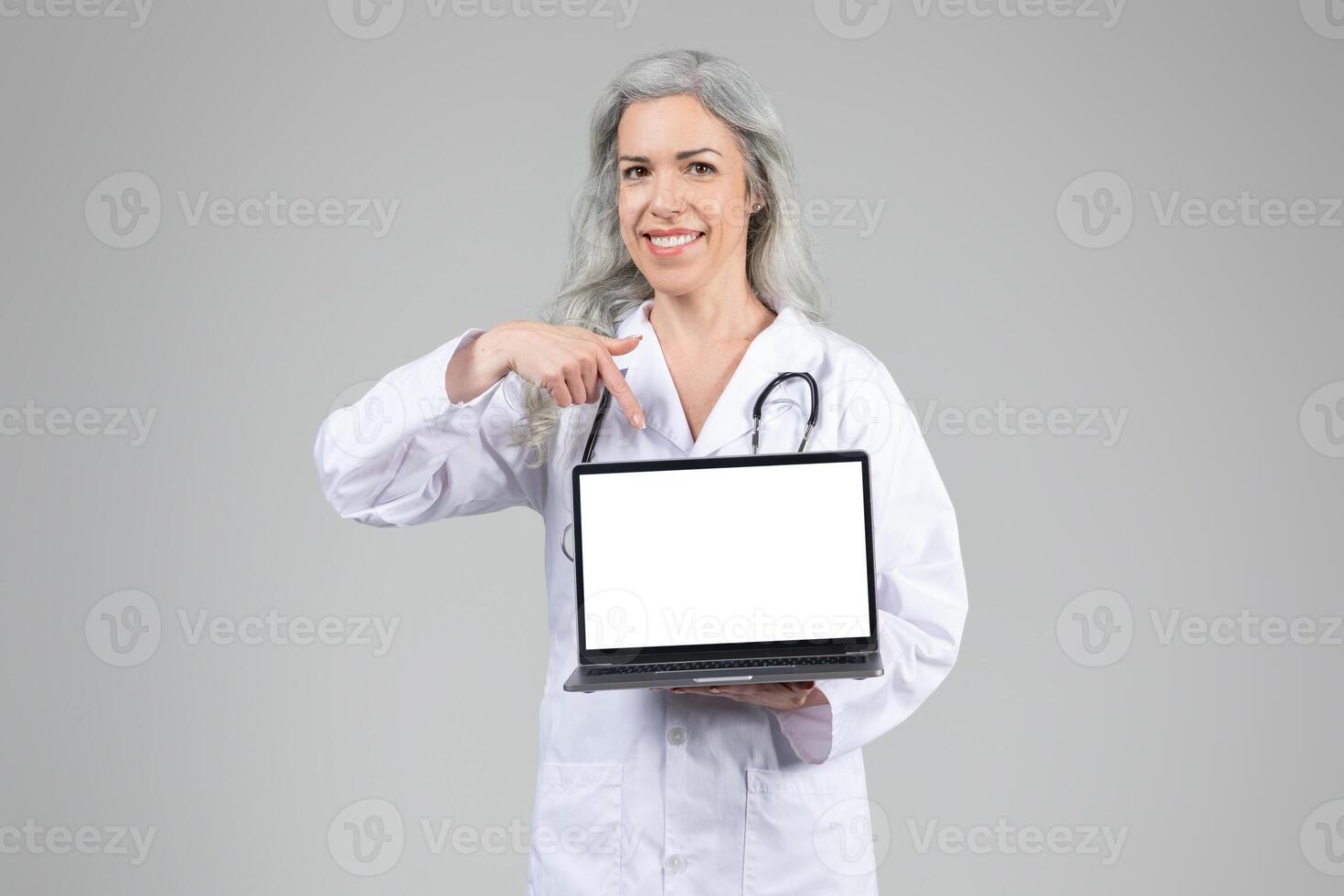 A middle aged woman wearing a white coat and stethoscope stands against a neutral background, smiling and pointing at a blank laptop screen, representing modern healthcare and telemedicine. photo