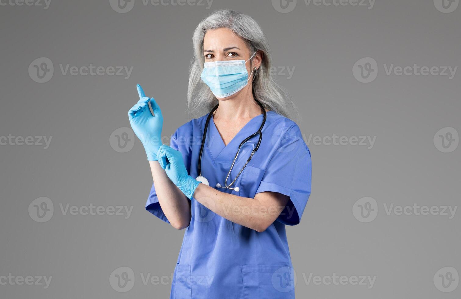 A middle aged woman in scrubs wears a mask and gloves while preparing for a medical examination. She appears focused and ready to provide healthcare in a clinical setting. photo