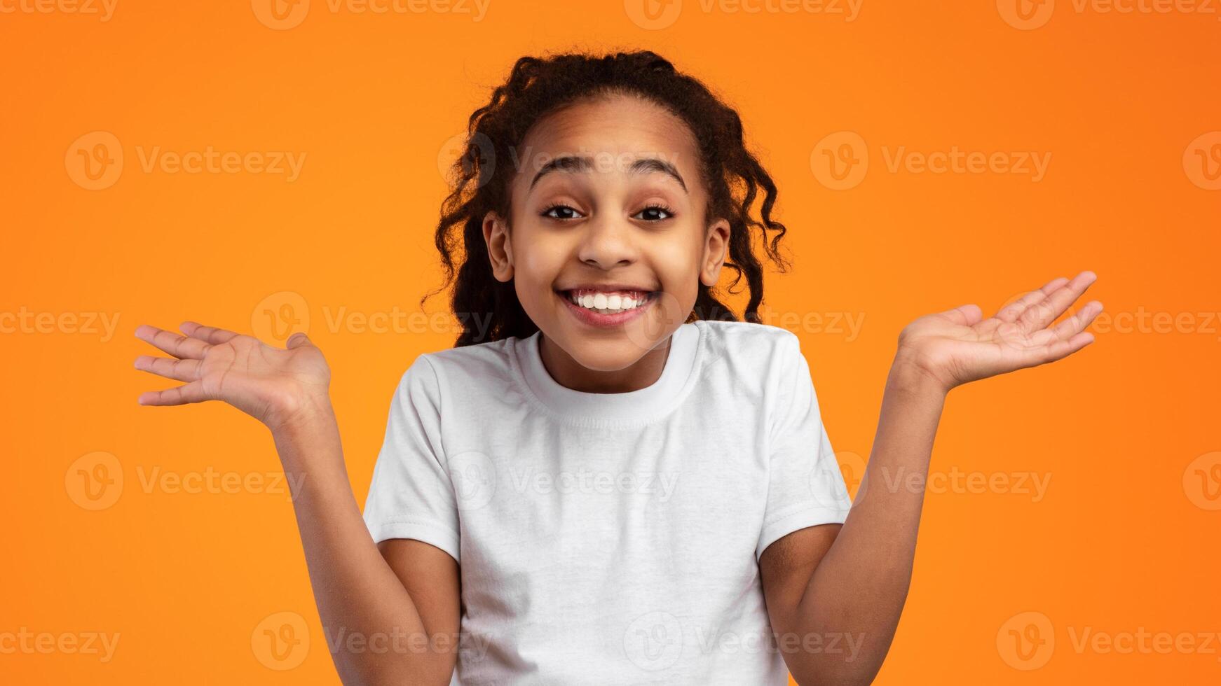 Studio shot of happy positive emotional young black girl spreading palms raising hands up and looking at camera. Smiling kid seeing something wonderful isolated over yellow studio background photo
