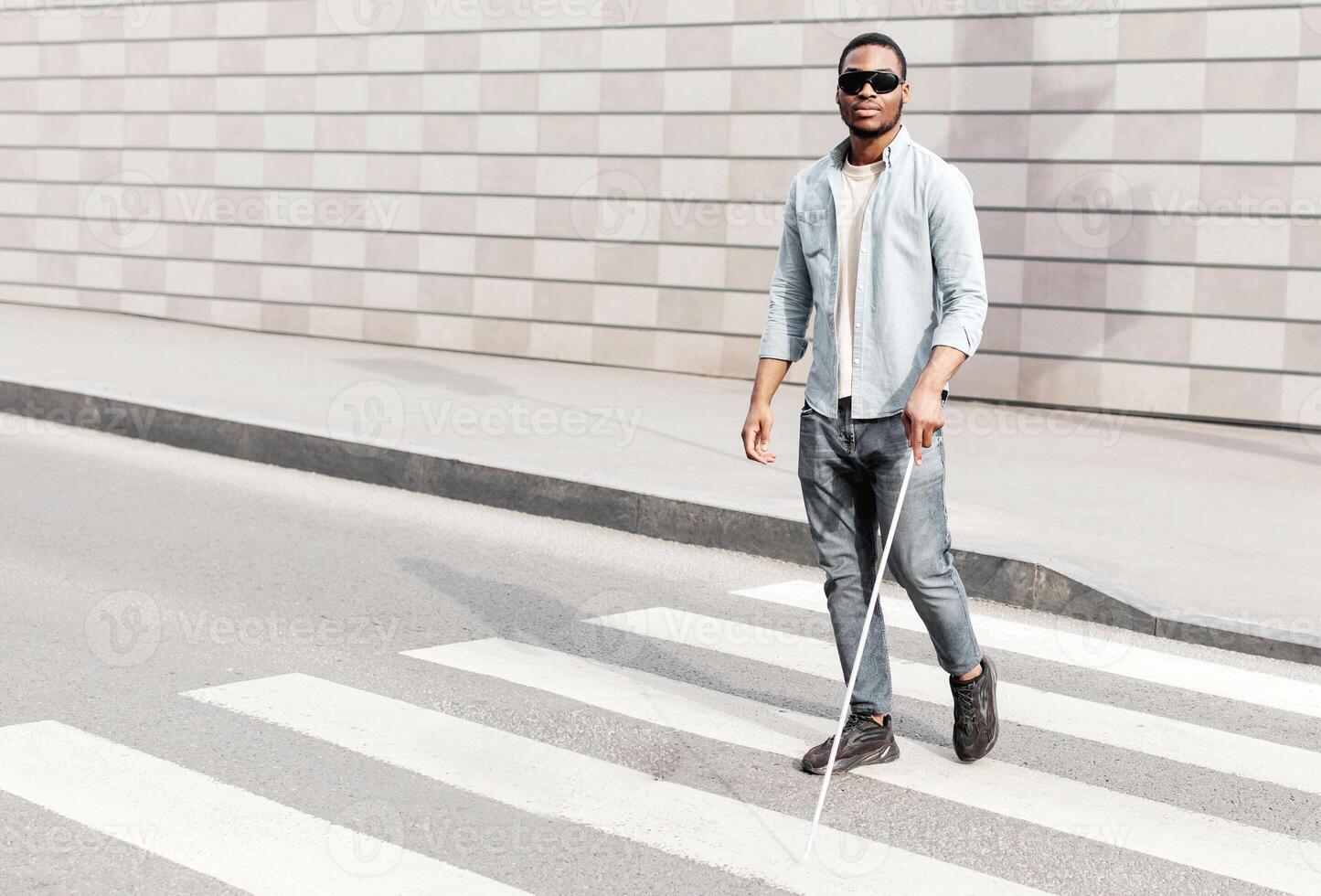Full length of young black man with visual disability wearing dark glasses, using walking cane to cross street at city centre, copy space. Impairment and urban lifestyle concept photo
