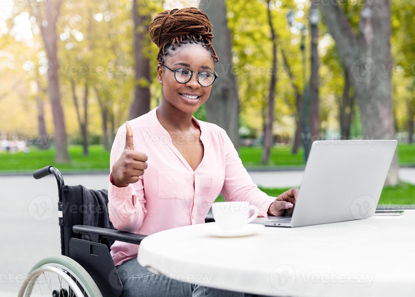 Joyful disabled black lady in wheelchair using laptop pc, working online, showing thumb up gesture, recommending remote job for handicapped people at autumn park, copy space photo