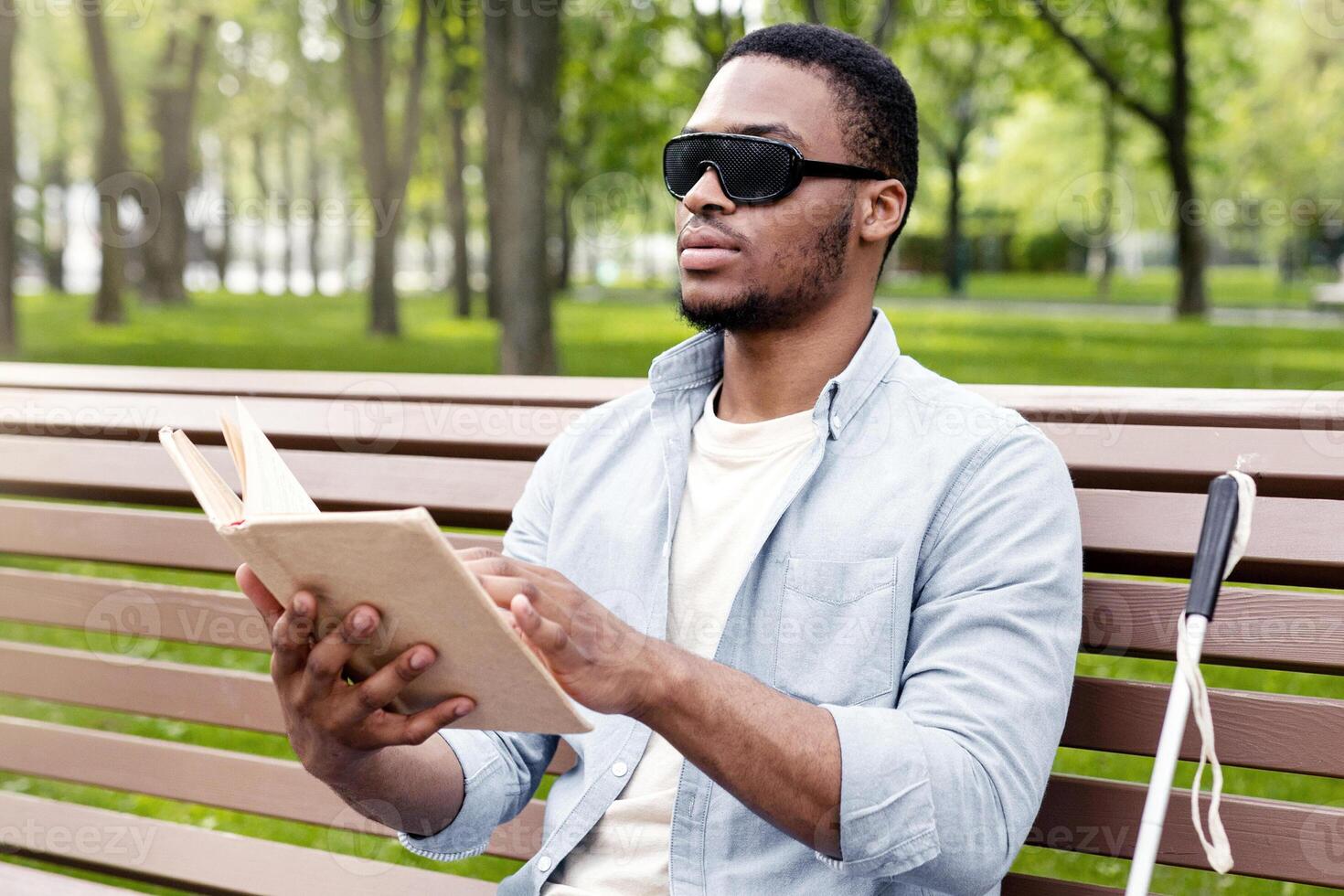 Young black visually impaired man sitting on bench in city park, reading Braille book outdoors. Millennial blind guy in dark glasses studying or enjoying entertainment literature, outside photo