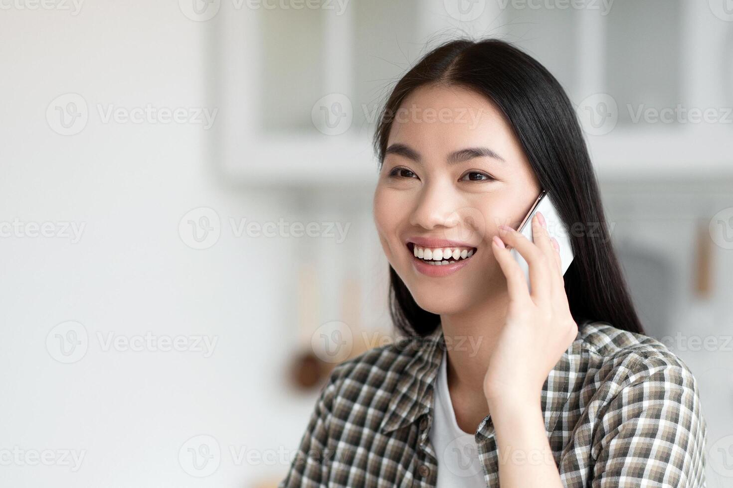 Beautiful young asian lady having healthy breakfast and talking on cellphone, smiling and looking at copy space. Pretty woman sitting at kitchen, eating oatmeal and calling girlfriend or lover photo