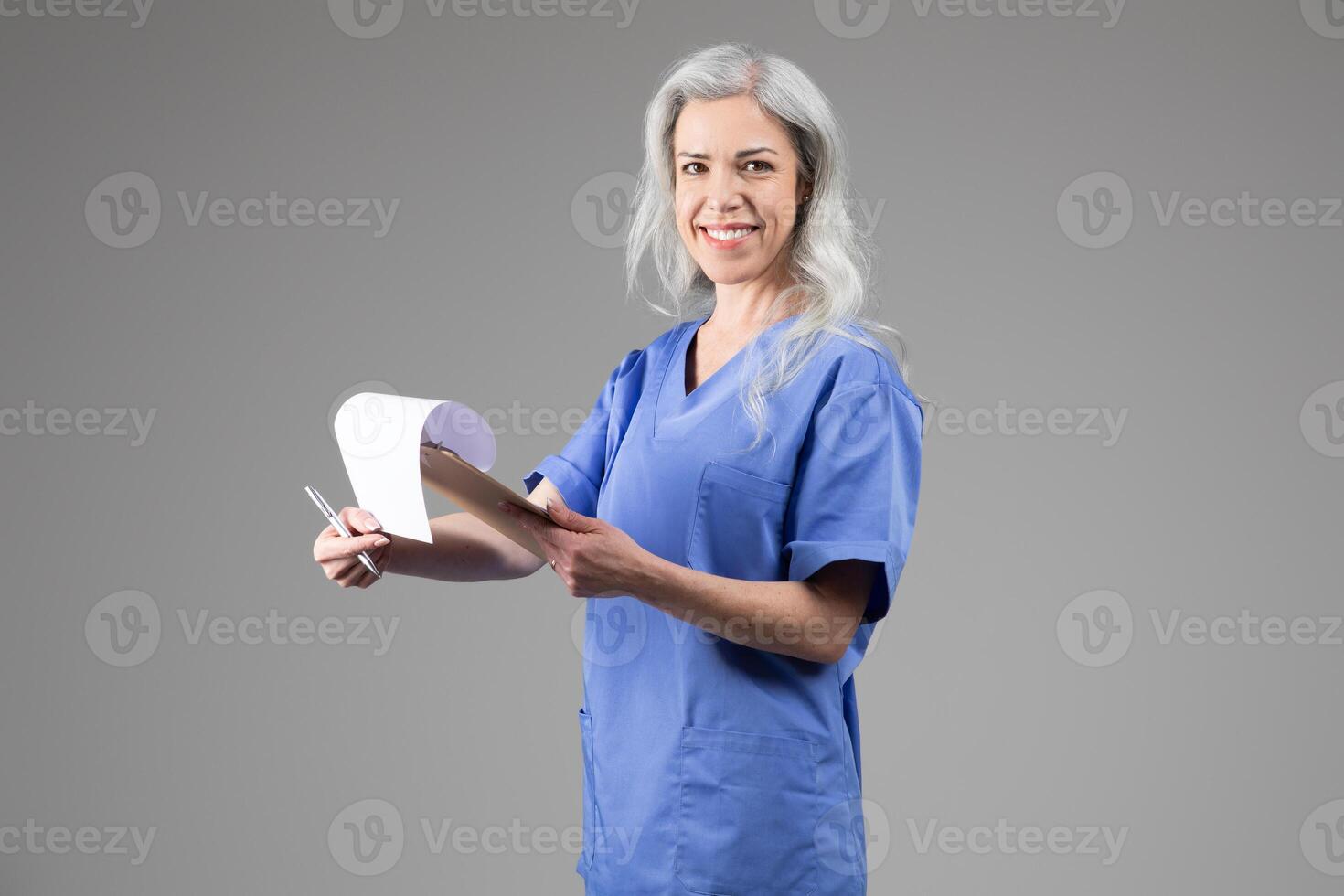 A middle aged woman dressed in blue scrubs is smiling as she holds a patient chart while standing against a neutral background. Her confident demeanor reflects her dedication to healthcare. photo