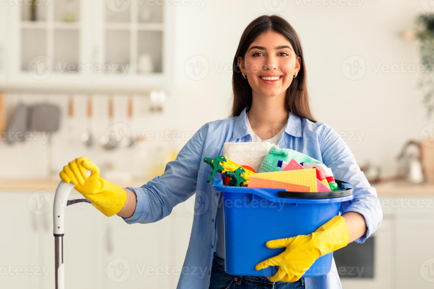 Chores. Portrait of cheerful young housewife holding bucket with cleaning supplies and mop, smiling and looking at camera standing in kitchen. Professional cleaning service specialist offering help photo