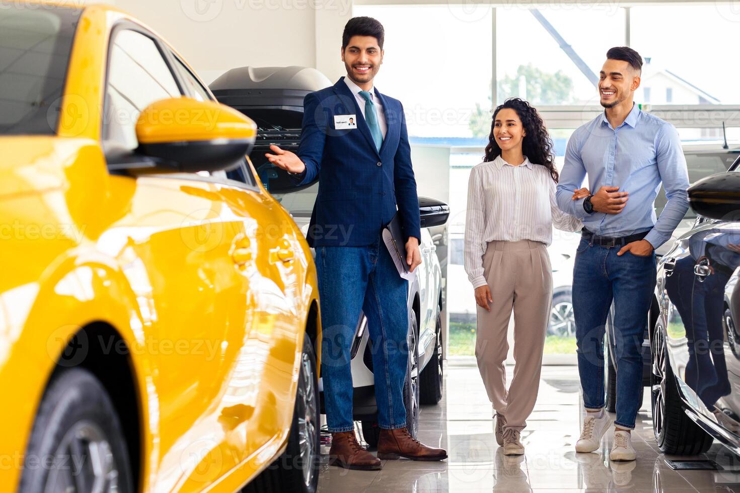 Wealthy middle-eastern young couple having conversation with good-looking sales assistant at auto showroom, looking at nice yellow car and smiling, full length photo, panorama photo