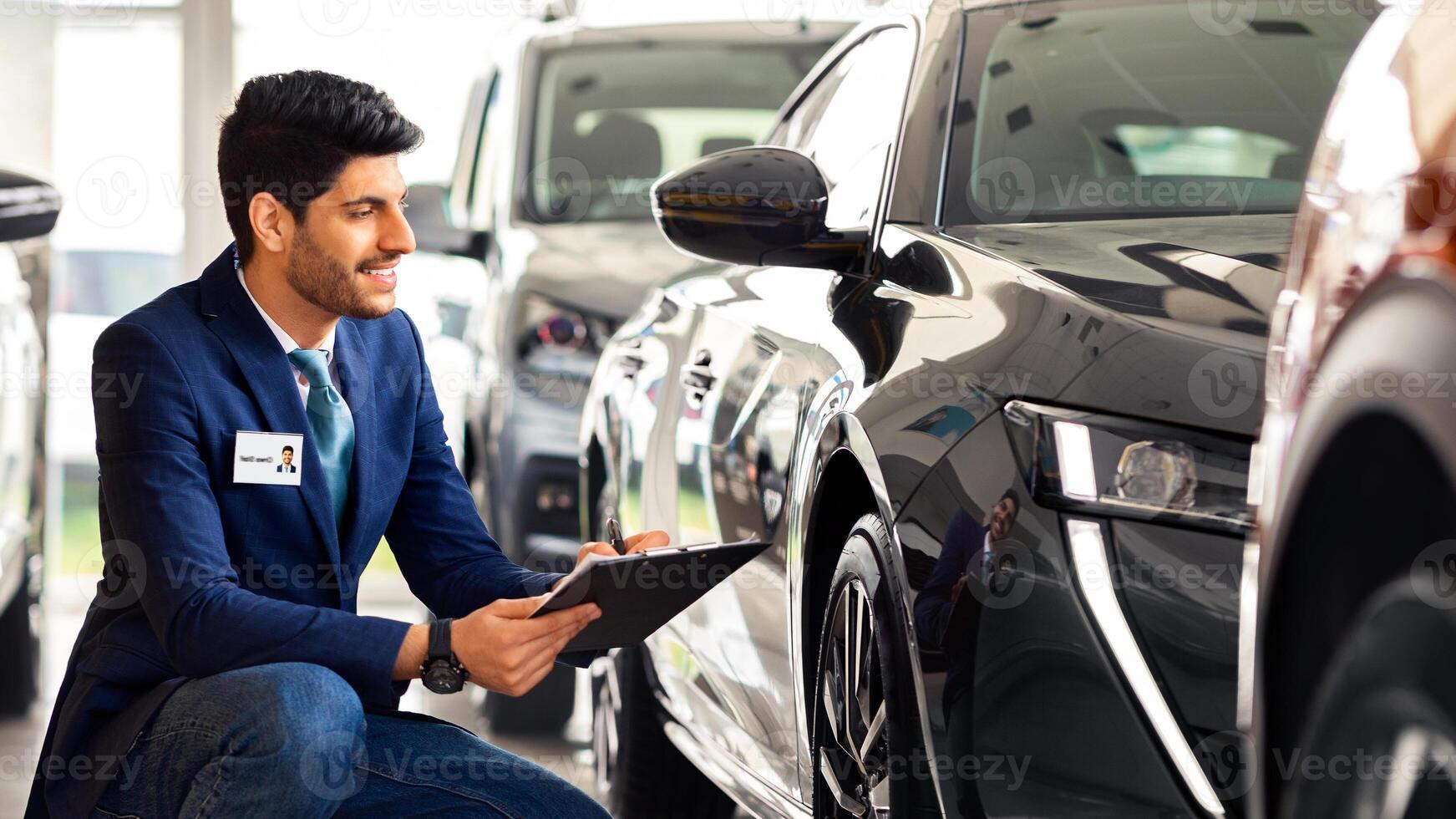 Middle-eastern guy sales manager in luxury car salon making checkup, holding chart and taking notes while looking at new comming automobile surface, side view, panorama with copy space photo