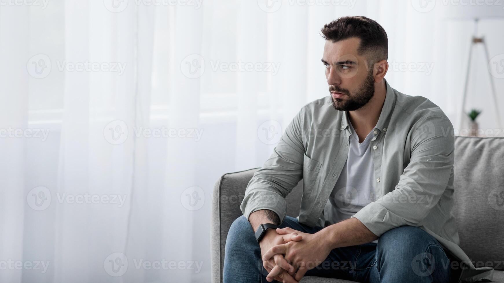 Thoughtful serious bearded millennial male sit on sofa alone at home, lost in thoughts, thinking about problem solving, feeling lonely, make important decision, have psychological problem, side view photo