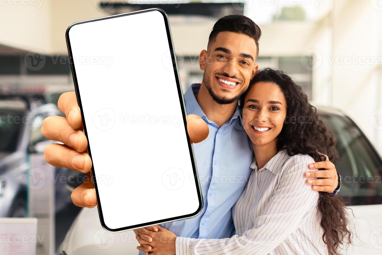 Loving millennial muslim family husband and wife embracing and showing newest smartphone with white blank screen, recommending mobile application for auto dealership, buying car at showroom photo