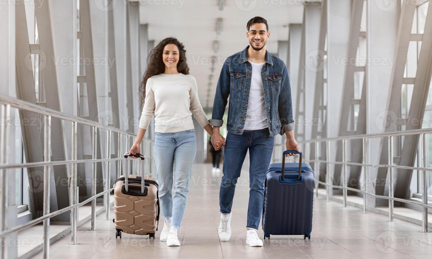 Time For Trip. Cheerful Arabic Couple Walking With Luggage At Airport Terminal, Smiling Middle Eastern Spouses Carrying Suitcases While Going To Flight Departure Gate, Looking At Camera, Copy Space photo