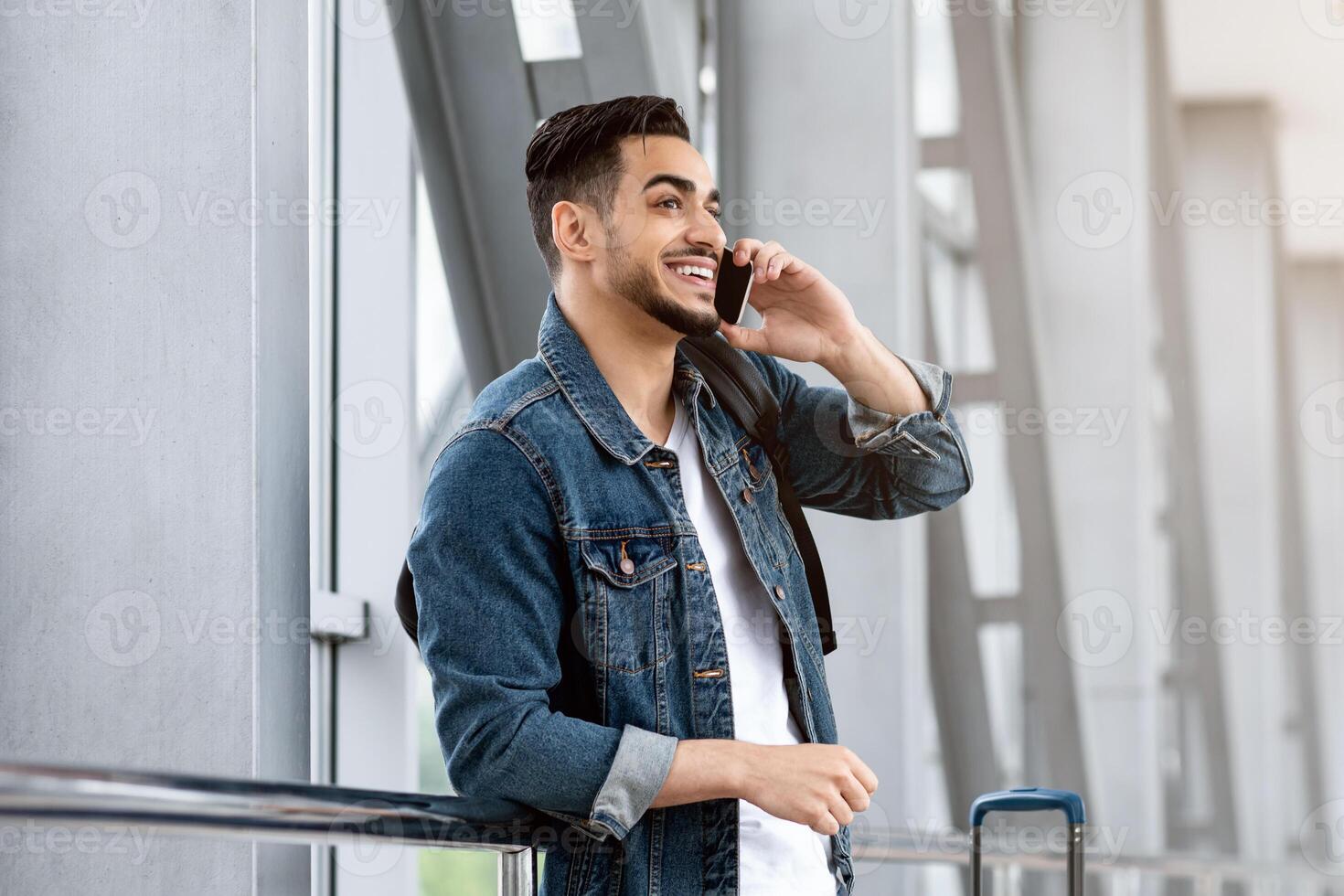 Mobile Communication. Smiling Young Arab Guy Talking On Cellphone At Airport, Millennial Middle Eastern Man Standing With Luggage Near Window At Terminal And Enjoying Pleasant Call, Copy Space photo
