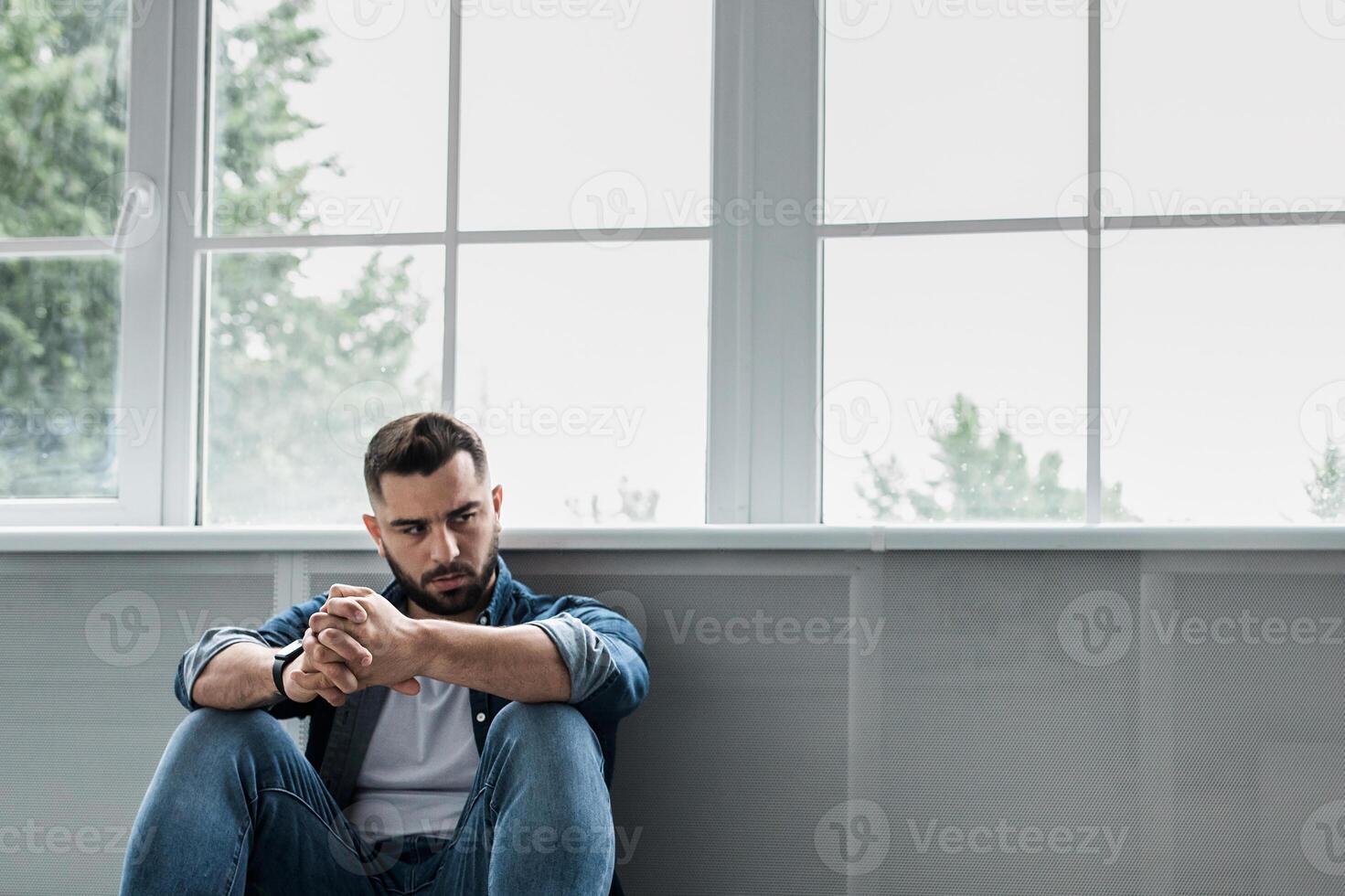 Man feeling sad, tired and worried, suffering depression in mental health, problems and broken heart. Frustrated young handsome bearded guy sitting on floor near large window, copy space, panorama photo