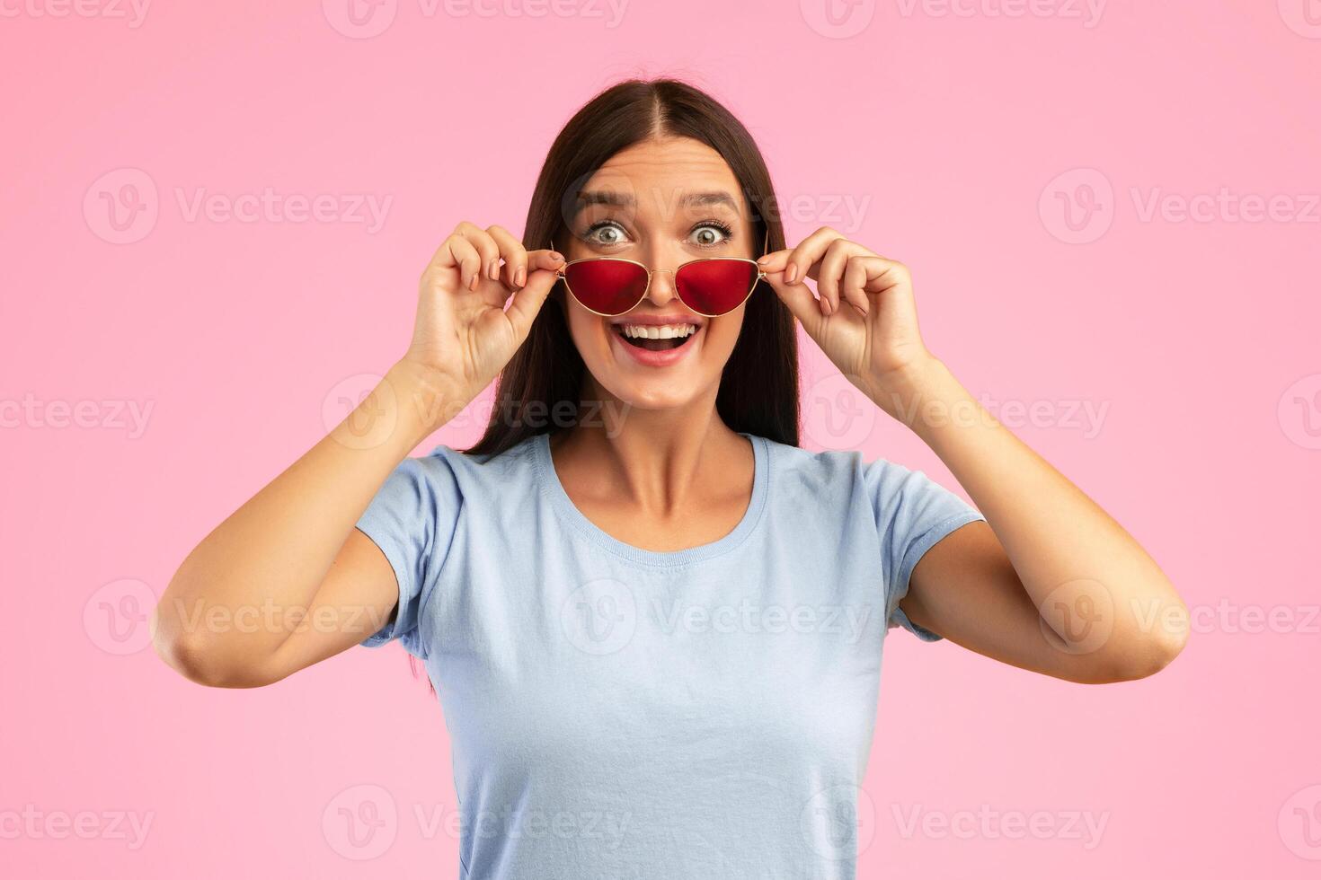 Wow. Portrait of trendy woman posing at studio, taking off red sunglasses, looking at camera with surprised facial expression isolated on yellow background. It's Unbelievable. Summer Sale photo