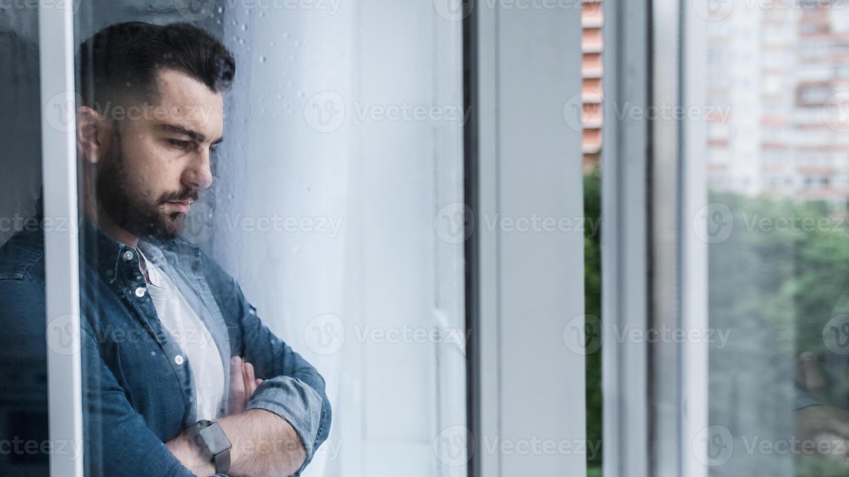 Depressed caucasian man losing job and heartbroken, alone near window. PTSD, mental health and depression. Sad unhappy young handsome guy with crossed arms looking at wet window in kitchen interior photo