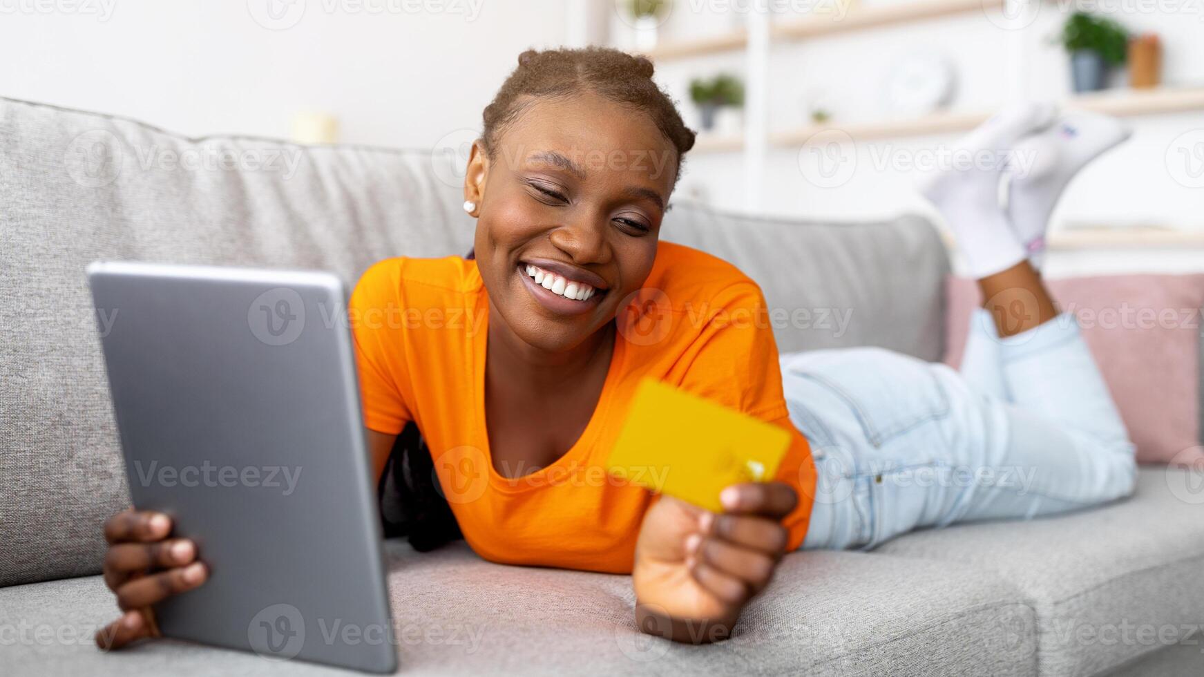 Happy black lady lying on sofa with tablet pc and credit card, shopping on internet at home. Positive African American woman purchasing stuff online in web store, full length portrait photo