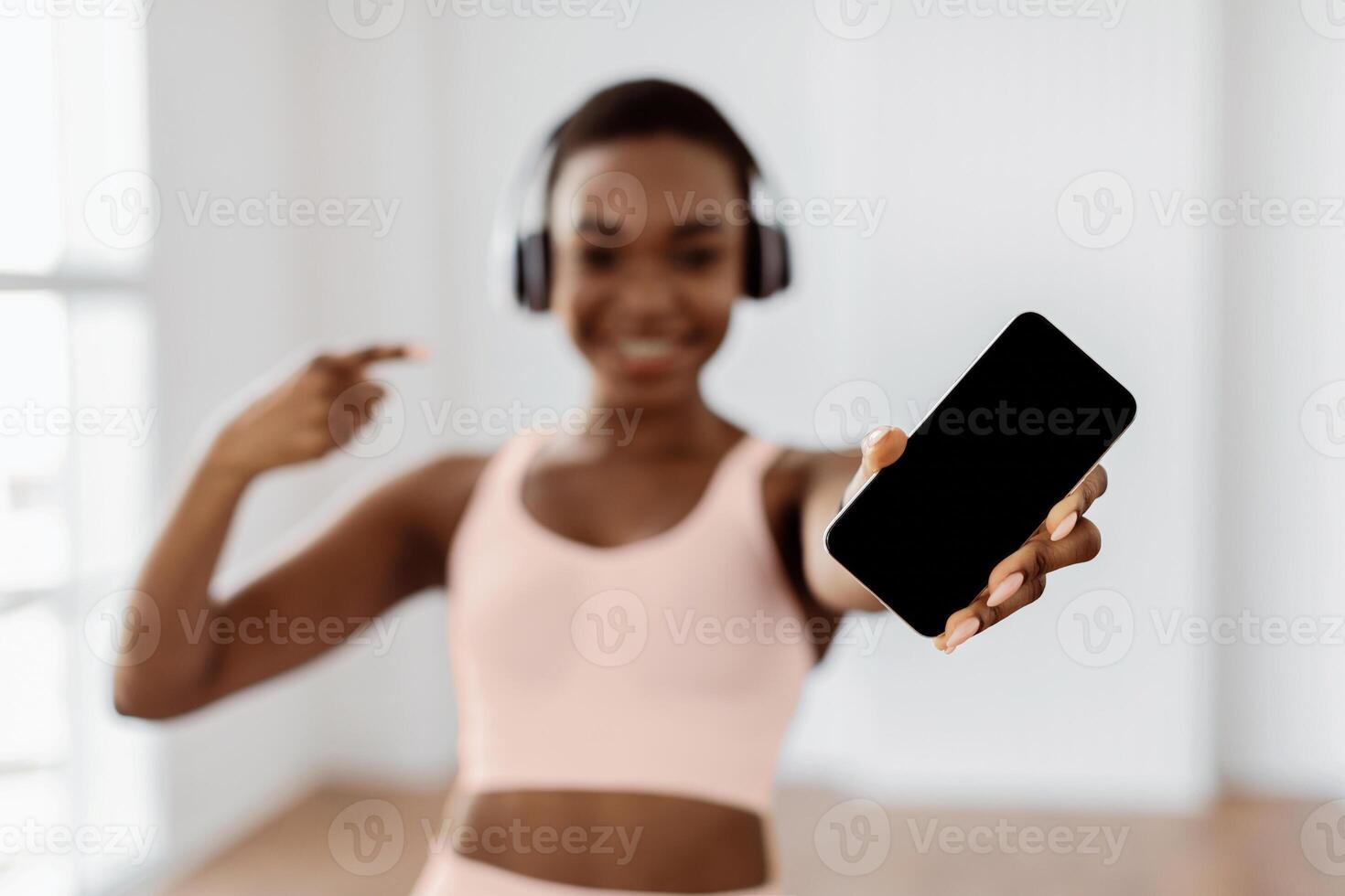 Sports App Concept. Fit young African lady holding and showing smartphone with sporty empty screen for mock up template, pointing at isolated gadget display, blurred background, selective focus photo