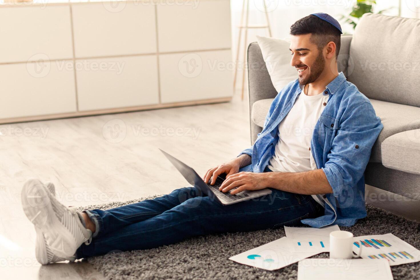 Successful Career And Distance Job Concept. Portrait Of Smiling Israeli Man In Kipa Working On Laptop Sitting On Floor Carpet With Papers, Leaning On Sofa At Home Office, Modern Interior, Free Space photo