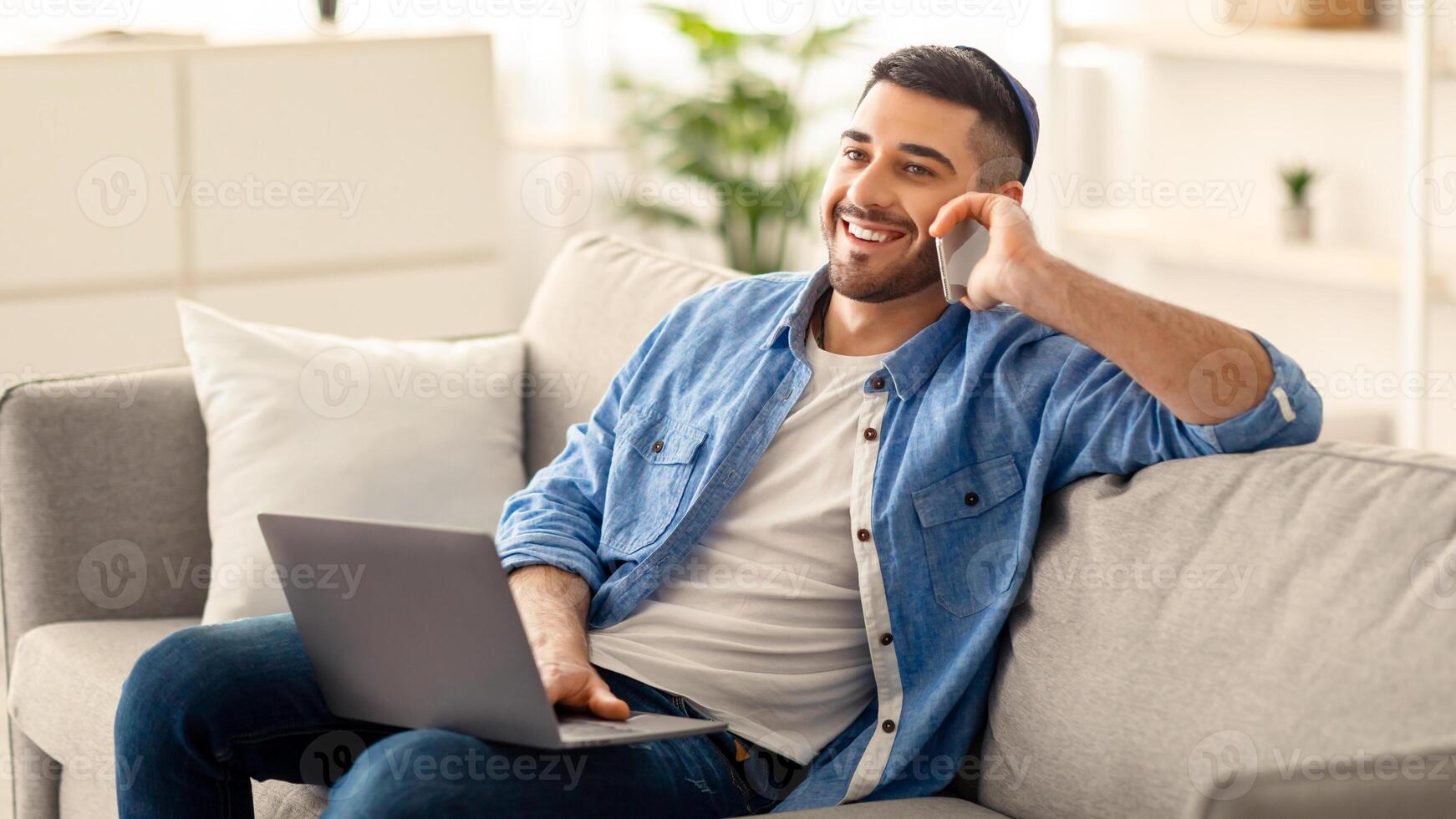 Portrait of smiling young jew man in yarmulke talking on mobile phone, working remotely sitting on comfortable couch with laptop at home office. Positive guy making business call, blurred background photo